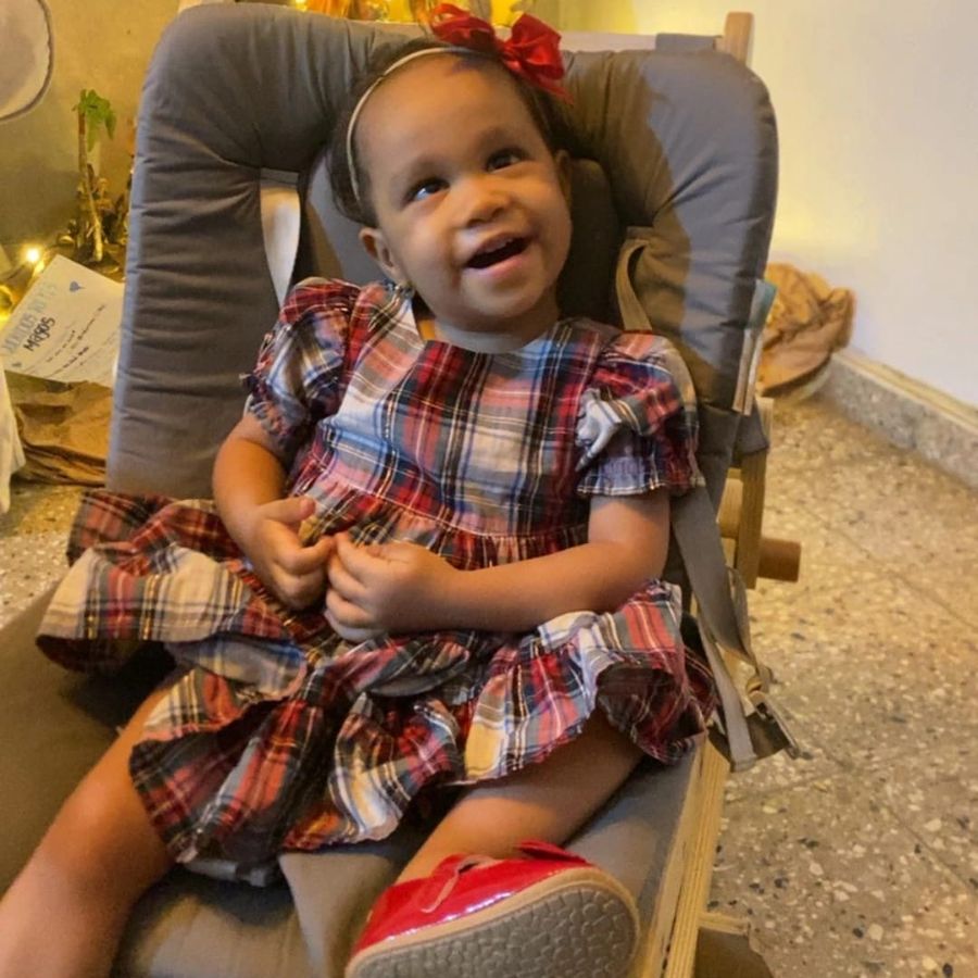 Carmen, a smiling two-year-old girl in a plaid dress, sitting in her chair